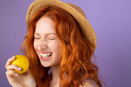 Cheerful widely smiling redhead woman wearing a straw hat holding an apple in her hand and posing over violet background.の写真素材
