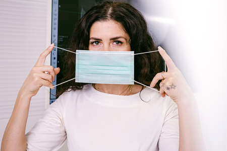 Close up photo of a brunette woman in white holding medical mask covering her face looking at the camera.の写真素材