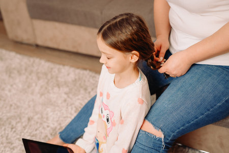 Cropped photo of mother in jeans sitting on the sofa and weaving braids for her daughter at home, girl sitting with closed eyes.の写真素材