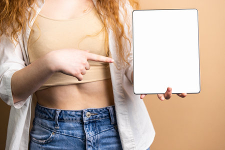 Cropped photo of a young woman with long red hair pointing with index finger at the tablet with white screen keeping in another hand.の写真素材