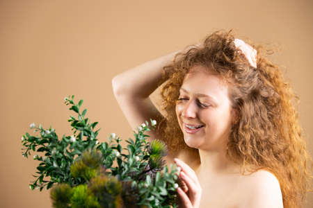 Photo of pretty redhead curly lady with big bunch of green flowers touching them with the hand and looking with smile on her face.の写真素材