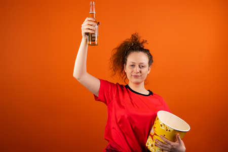 Relaxed fan football woman wearing red uniform with a bowl of popcorn and keeping a bottle of beer raising hand up.の写真素材