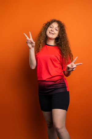 Full-length photo of a fan footbal redhead woman in team uniform keeping hands up with two fingers...