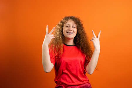 Joyful pretty redhead smiling woman in red uniform of her football team raising hands up holding fingers as a sign of victory.の写真素材