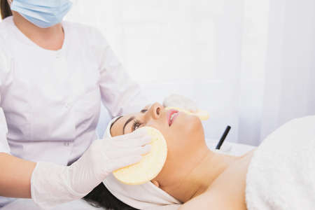 Beautician in disposable medical mask and gloves cleanses skin of beautiful woman with a sponge, spa treatment skincare face. Cleansing and tonifyingの写真素材