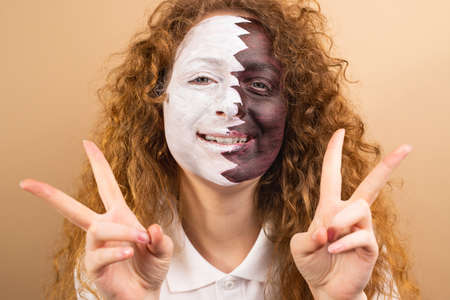 Headshot of optimistic fan football redhead woman with face painted in Qatar flag showing victory sign on yellow background.の写真素材