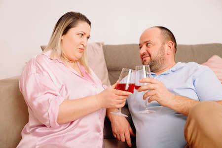 Happy caucasian couple relax on cozy sofa at home resting holding in their hands two glasses of wine and looking at each other with love.の写真素材