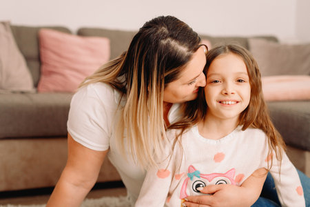 Close up photo of two people mother and her little nice daughter sitting on the carpet at home and mother hugging and kissing her little girl on the cheek.の写真素材