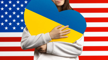 A girl holds a heart to her chest with the flag of Ukraine, and behind her the flag of the United States of America.の写真素材