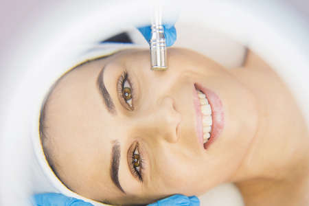 Close up photo of a beautiful smiling woman face looking at the camera through the magnifying glass and enjoying the rejuvenation procedure in a beauty salon.の写真素材