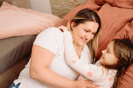 Daughter and mother lying down on big pillow hugging each other with smiling facial expression, family time together.の写真素材