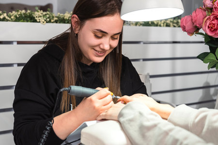 Master professional in beauty salon in black uniform using electric nail file drill making manicure to a woman client.の写真素材