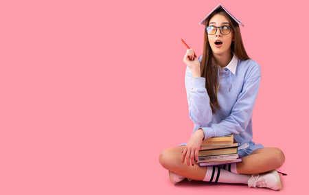 Photo of amazing young and beutiful school girl sitting isolated over pink wall background, looking aside and showing copyspace pointing, product placement.の写真素材