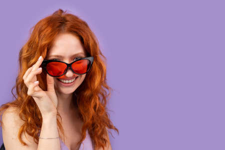 Young redhead woman in spring summer dress smling widely taking off her red sunglasses on purple background and copy space for advertising.の写真素材
