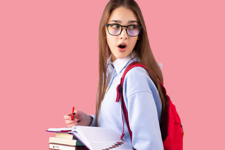 Amazing school girl in uniform holding a stack of books and a pen looking over her shoulder with open-eyed expression.の写真素材