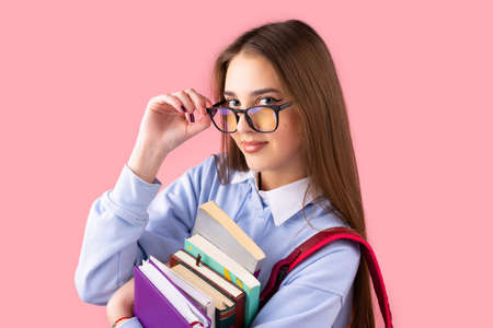 Pretty girl teen student with backpack holds books touching glasses with fingers having smart facial expression, education in high school concept.の写真素材