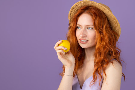Beauty portrait of a young attractive redhead girl with curly hair wearing a straw hat with an apple in her hand on purple background and copy space.の写真素材