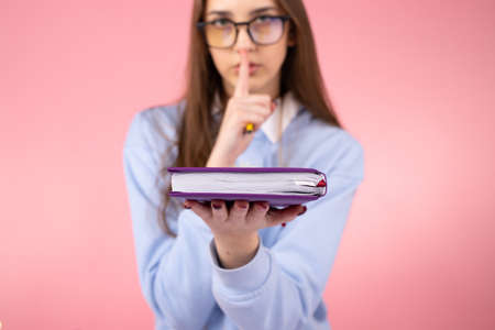 Blurry photo of a student pretty girl wearing glasses in uniform making silence sign with an index figer and holding a book in her hand.の写真素材