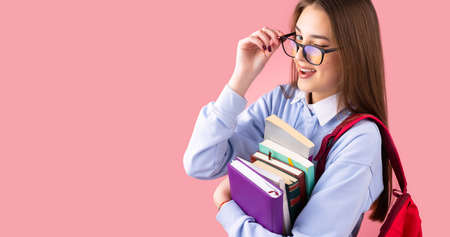 Side view photo of a school teenage girl in uniform with backpack and books giving a wink pushing back glasses, college concept.の写真素材