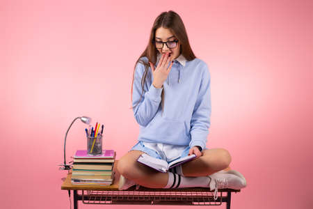 Amazing pretty beautiful school girl in blue uniform sitting on the table reading a book and covering her mouth with a hand.の写真素材