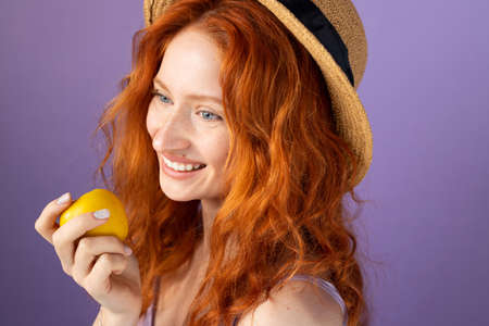 Redhead girl in a straw hat smiling widely holding an apple close to her face on purple background, health nutrition concept.の写真素材
