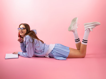 Satisfied beautiful student girl wearing blue uniform and glasses lying on the floor on her stomach and raised her legs up bending her knees and reading the book.の写真素材