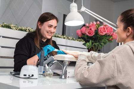 Young manicurist wearing gloves with joy looking at a client while removing an old manicure from a young woman with a nail file in a beauty salon.の写真素材