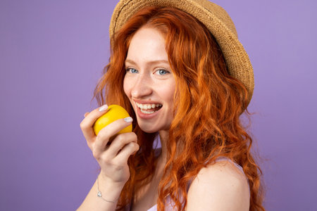 Young redhead woman with blue eyes wearing a straw hat smiling widely holding an apple ready to eat on purple background.の写真素材