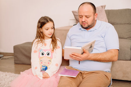 Father and daughter spending time together, reading a book while sitting together on the floor at home, family staying at home concept.の写真素材