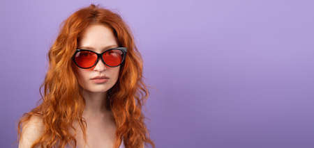 Pretty young woman with red hair wearing sunglasses for the summer time posing on purple background with copy space.の写真素材