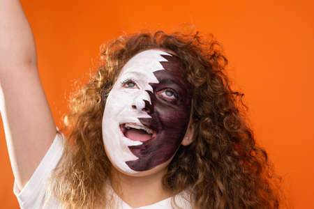 Pretty young redhead woman football supporter with opened mouth and raised hand up, painted face in Qatar flag, fan supporter concept.の写真素材