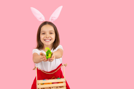 Cheerful little girl widely smiling wearing bunny ears on head in dress with basket ful of painted eggs in front of her posing on pink background and copy space.の写真素材
