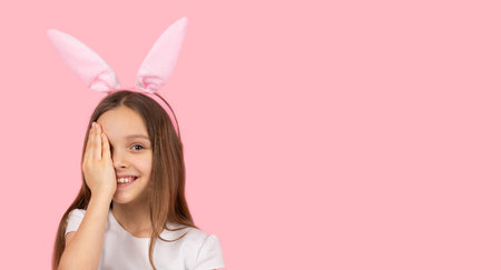 Picture of a blond little girl wearing bunny ears on head smiling for the camera and covering one eye with a palm on pink background and copy space.の写真素材