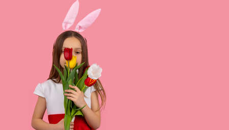 Portrait of good looking girl wearing bunny ears on head in a beautiful dress hiding her face with a bunch of tulips on pink background and copy space.の写真素材