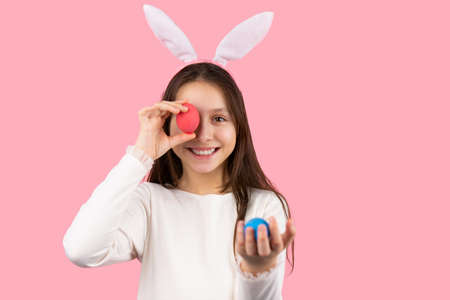 Close up photo of a cheerful girl with bunny ears on her head posing with one egg holding in her hand and with another egg covering one eye.の写真素材
