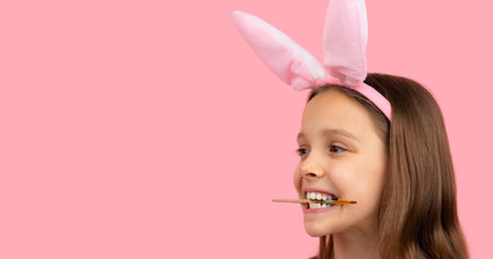 A cheerful girl with rabbit ears on her head keeping a brush with her teeth for painting eggs posing on pink background and copy space.の写真素材