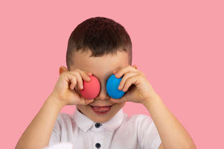 Little boy in white t-shirt covering his eyas with two egg red and blue color and showing tongue posing on pink background.の写真素材