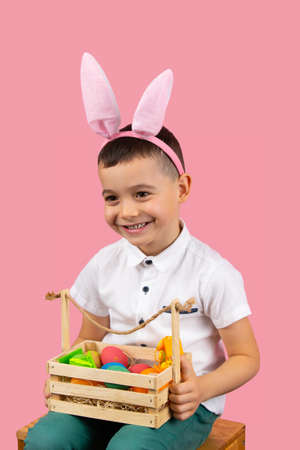 Cheerful boy sitting on a chair wearing rabbit ears holding basket with eggs on his knees posing on pink background.の写真素材