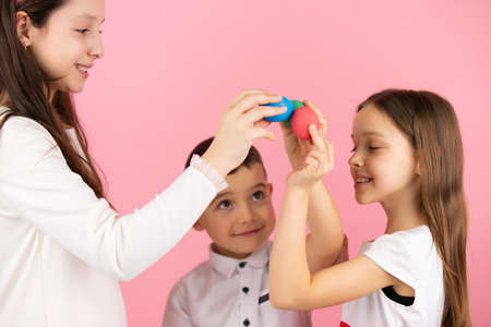 Family of three children holding their hands up with painted eggs and keeping them together on pink background, Easter concept.の写真素材