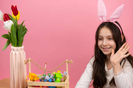 Cute little girl wearing bunny ears on head sitting at the table with a basket full of paints and vase with flowers waving to the camera.の写真素材