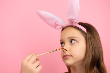 Cropped photo of a blond little girl wearing bunny ears looking aside and one hand painting her nose using brush.の写真素材