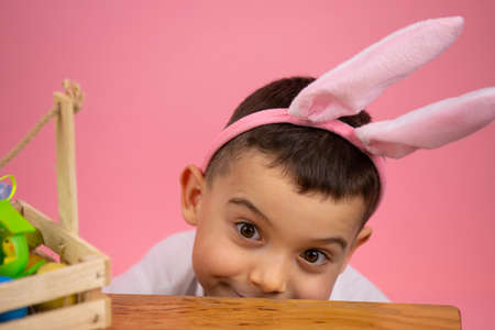 Pretty boy with bunny ears on head and widely opened eyes posing on pink background with a basket on the table.の写真素材