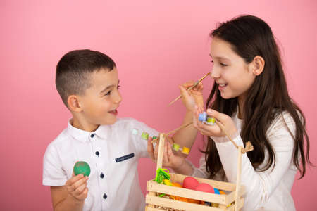 Happy family preparing for Easter, cute brother holding a green egg in his hand and using a brush painting his sister nose while she holding a basket with eggs.の写真素材