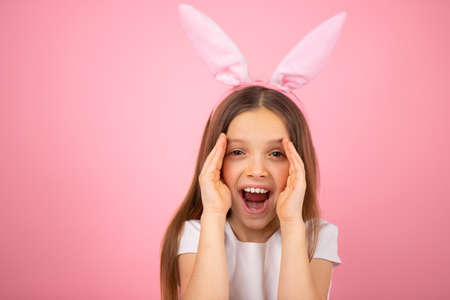 Close up photo of a little cute girl wearing bunny ears on head looking at the camera with widely opened mouth and covering her face on two sides with her palms on pink background.の写真素材
