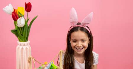 Close up photo of a happy pretty girl with bunny ears on head and a basket with paints on the table and a vase with flowers, Easter concept.の写真素材