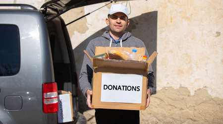 Full-length photo of a volunteer young man keeping in his hands a box with food for ukranian refugees, humanitarian aid concept.の写真素材