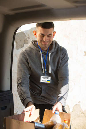Volunteer man with smiling facial expression taking the box of food from the car for the ukranian refugees running away from war.の写真素材
