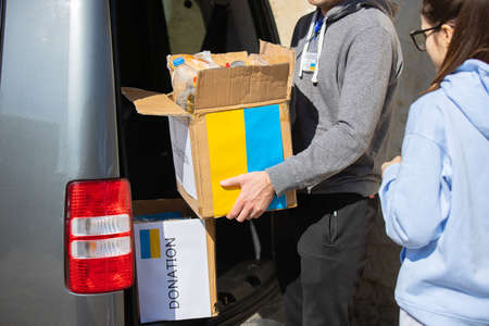 Two volunteers young woman and man helping refugees from ukraine, man taking out of the car a box painted in ukranian flag with food.の写真素材