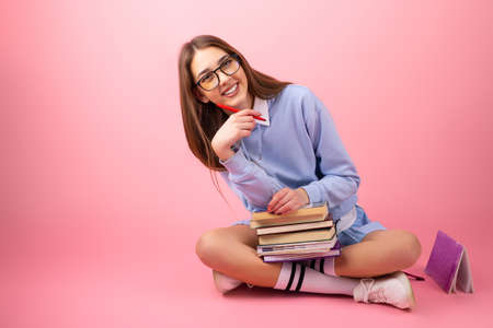 Joyful school girl with eyeglasses sitting on the floor with crossed legs and keeping a stack of books and a pen holding in her hand on pink background.の写真素材