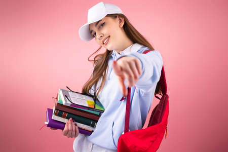Young smiling happy with backpack holds books and backpack with a cap on her head isolated stretched out her hand to the camera.の写真素材